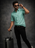 Man in a teal shirt and black pants holding sunglasses and a suitcase against a dark background