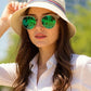 Woman wearing a white shirt, green sunglasses, and a beige hat with a blurred cityscape background